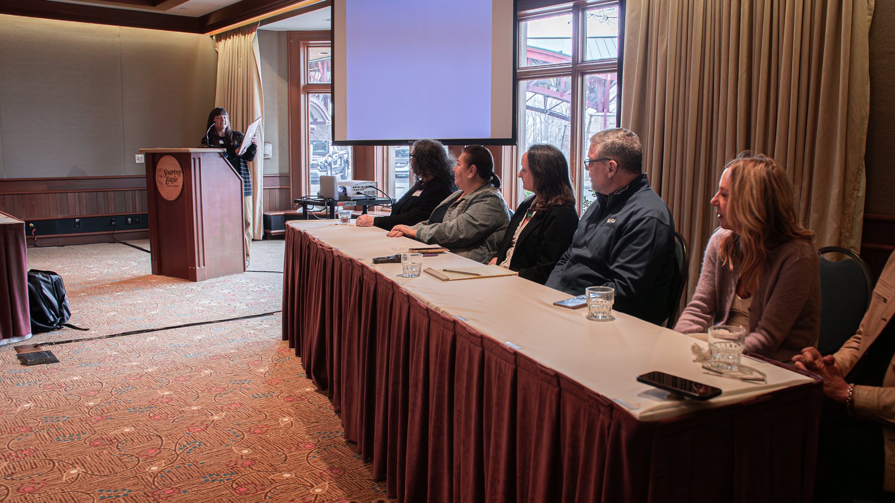 A speaker at a podium addresses a panel of Tribal and state housing leaders at a NAIHC roundtable